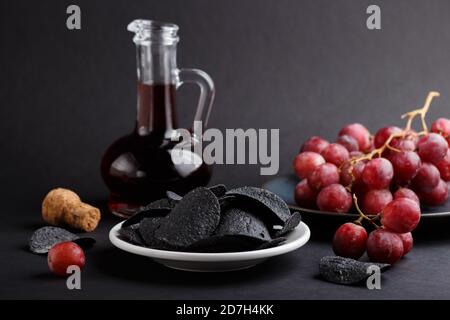 Patatine nere con carbone, aceto balsamico in vetro, uva rossa su un piatto di ceramica blu su fondo nero. Vista laterale, primo piano, selettivo Foto Stock