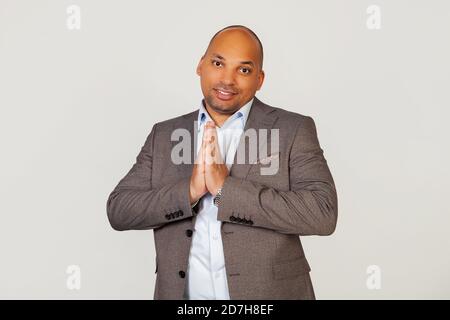 Ritratto di un giovane uomo d'affari afro-americano sorridente, palma insieme davanti a lui, tenendo le mani in un gesto di preghiera, chiedendo perdono per un errore. In piedi su uno sfondo grigio Foto Stock