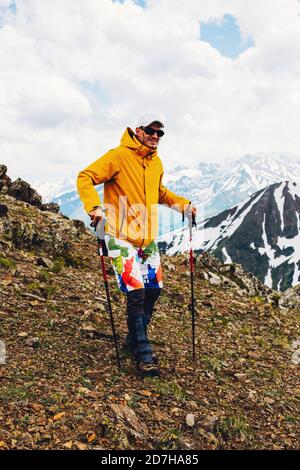 Giovane turista maschile vestito in abiti turistici e camminando lungo la cima di una montagna sullo sfondo di catene montuose innevate. Mountai Foto Stock