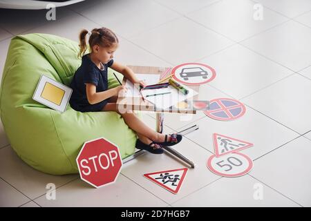 Carino bambina si siede sulla sedia verde morbido vicino al tavolo con matita e fogli di carta. Vicino all'automobile moderna e ai cartelli stradali sul pavimento Foto Stock