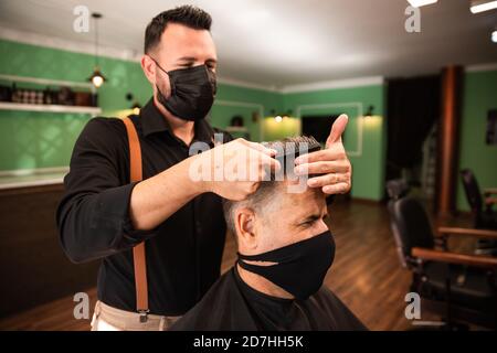 in un barbiere un barbiere combe un uomo cresciuto con la mano e il pettine, indossano maschere per la prevenzione del coronavirus pandemico. il cliente è seduto. anche Foto Stock