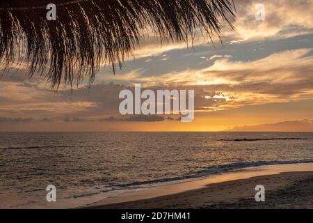 Viste da sotto un ombrello di paglia dello spettacolare tramonto a Playa del Duque con l'oceano calmo, la spiaggia vuota, il cielo nuvoloso colorato e l'isola di la Gomera Foto Stock