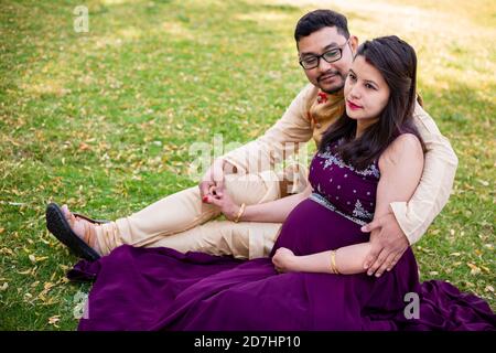 Giovane donna indiana asiatica incinta con suo marito in seduta rilassante nel parco o in giardino, giovani genitori che guardano la macchina fotografica aspettandosi il bambino. Foto Stock