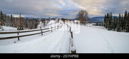 Campagna secondaria strada alpina in remoto villaggio di montagna, nevicate e recinzione in legno sul lato del percorso Foto Stock