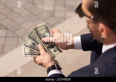 Primo piano di riuscito imprenditore americano considerando wad di banconote da 100 dollari mentre si siede sul marciapiede. Vista posteriore Foto Stock