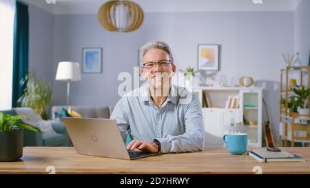 Bell'uomo di mezza età usa il computer portatile e sorride sulla fotocamera. È nel salotto accogliente ed elegante. Felice uomo di successo funziona sul computer Foto Stock