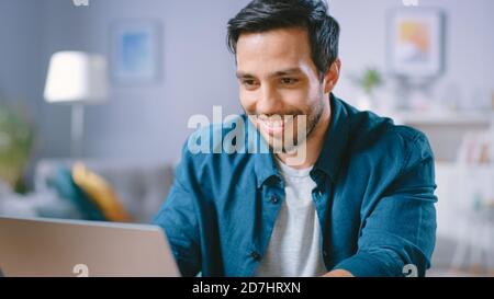 Ritratto di bell'uomo giovane che usa il computer portatile a casa, guardando e facendo un po' di laughing ai contenuti. Happy Hispanic Man funziona su computer in una vita accogliente Foto Stock