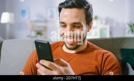 Happy Young Man utilizza lo smartphone mentre si è seduti su un divano a casa. L'uomo naviga attraverso Internet, Watches Video e utilizza Social Networks a casa. Foto Stock