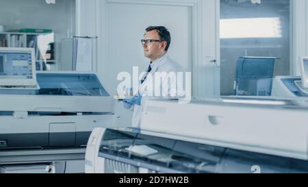 Il ricercatore maschile attraversa il laboratorio con un vassoio di provette riempito di campioni. In background Working Laboratory con innovativo Foto Stock