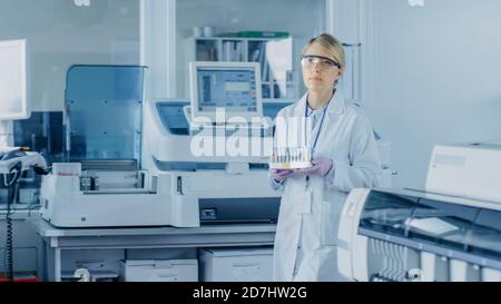 La scienziata di ricerca femminile attraversa il laboratorio con un vassoio di provette riempito di campioni. In background persone che lavorano in laboratorio con Foto Stock