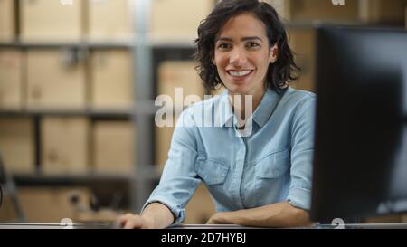 Il responsabile dell'inventario femminile lavora seduto alla sua scrivania, lavorando al computer e sorridendo alla videocamera. In background magazzino Storeroom con scaffali Foto Stock