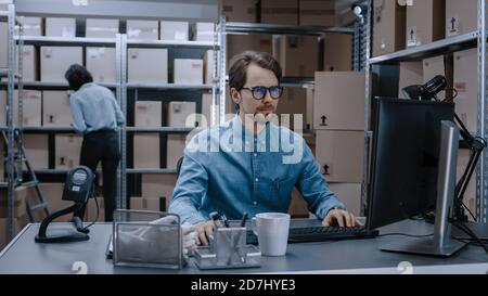 Warehouse Inventory Manager lavora su computer mentre si siede alla sua scrivania, in background, Female Worker Check Shelf per un pacchetto di consegna. Gli scaffali sono Foto Stock