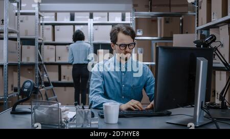 Warehouse Inventory Manager lavora su computer mentre si siede alla sua scrivania, in background, Female Worker Check Shelf per un pacchetto di consegna. Gli scaffali sono Foto Stock