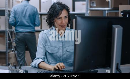 Warehouse Female Inventory Manager lavora su un computer mentre si siede alla sua scrivania, in background Male Worker utilizza Digital Tablet computer per controllare Foto Stock