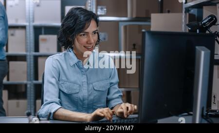 Magazzino Female Inventory Manager lavora su un computer mentre si siede alla sua scrivania, in background scaffali pieni di scatole di cartone pacchetti pronti per Foto Stock