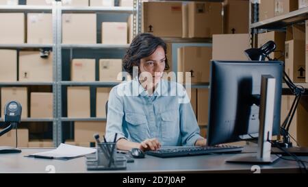 Il responsabile dell'inventario femminile lavora su un computer mentre si siede alla sua scrivania, marcare gli ordini nella lista di controllo degli Appunti. In background magazzino Storeroom Foto Stock