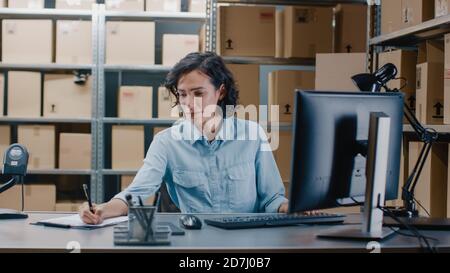 Il responsabile dell'inventario femminile lavora su un computer mentre si siede alla sua scrivania, marcare gli ordini nella lista di controllo degli Appunti. In background magazzino Storeroom Foto Stock