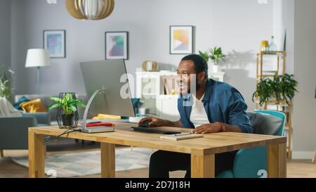 Young Black Man, scioccato e sorpreso, utilizza il Personal computer a casa. In background accogliente soggiorno. Foto Stock