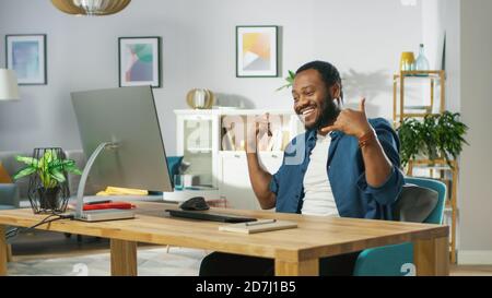 L'uomo giovane eccitato usa il calcolatore personale a casa, parla con l'amico e fa il gesto di 'segno di sacca. In background accogliente soggiorno. Foto Stock
