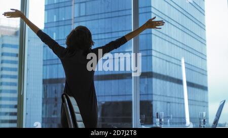 Indietro Vista della Businesswoman in piedi nel suo ufficio allungando, alzando le armi, celebrando il successo mentre guardando fuori dalla finestra con vista del paesaggio urbano. Foto Stock