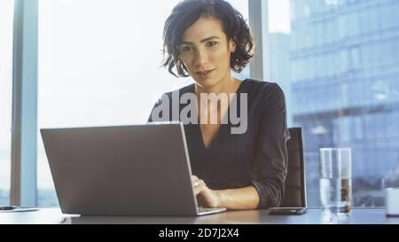 L'esecutivo femminile sicuro lavora su un laptop seduto alla sua scrivania in ufficio moderno con vista città di Grandiouse. Busiesswoman usa un laptop. Foto Stock
