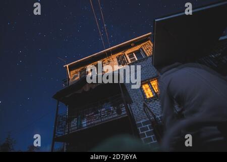 Foto notturna di una casa di campagna in mattoni con finestra illuminata solitaria con un cielo stellato su uno sfondo. Concetto di pace, magia, idilliaco, casa, sogni. Foto Stock