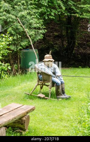 Scarecrow, fatto di fieno, vestiti e cappello, divertente uomo come figura, seduto su una sedia che tiene una canna da pesca vicino a uno stagno in Germania, Europa occidentale Foto Stock