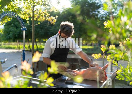 Chef maschile indossare maschera taglio carne di capra sul tavolo mentre in piedi in frutteto Foto Stock