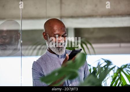 Sorridente freelance maturo che ascolta le chiamate tramite smartphone mentre appoggiandosi su una parete di vetro Foto Stock