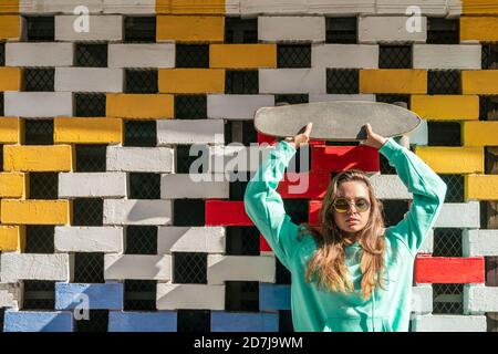 Giovane donna che solleva lo skateboard mentre si trova contro il muro di mattoni Foto Stock