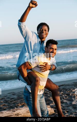 Giovane uomo che porta un amico piggyback sulla spiaggia durante la giornata di sole Foto Stock