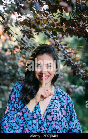 Primo piano di una giovane donna sorridente con un dito sulle labbra in piedi contro le piante nel parco Foto Stock