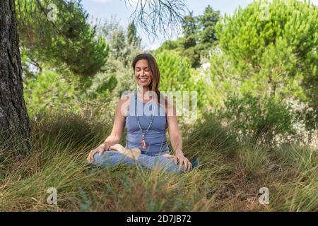 Donna sorridente che pratica yoga mentre si siede sotto l'albero sull'erba Foto Stock