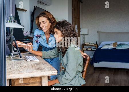 Madre che aiuta la figlia mentre usa il tablet digitale dal tavolo a. casa Foto Stock