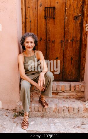 Donna matura seduta sulla porta contro la porta di legno Foto Stock