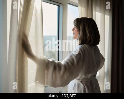 Donna anziana bionda che apre la tenda bianca mentre guardando attraverso la finestra in una camera d'hotel di lusso Foto Stock