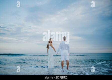 Donna che guarda dietro mentre tiene la mano dell'uomo in piedi dentro acqua in spiaggia Foto Stock