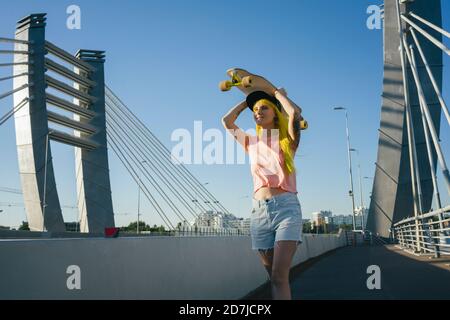 Giovane donna che solleva lo skateboard in testa mentre si sta in piedi sul ponte durante il giorno di sole Foto Stock