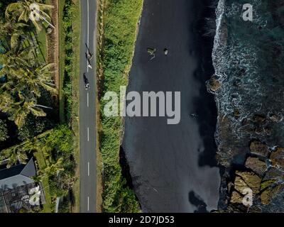 Indonesia, Bali, vista aerea dello skateboard maschile su strada asfaltata che si estende lungo la spiaggia costiera nera Foto Stock