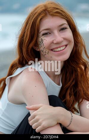 Donna sorridente con piccole pietre e sabbia sul viso seduta sulla spiaggia Foto Stock