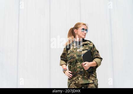 Soldato dell'esercito di biondo maturo che indossa occhiali da sole mentre tiene il digitale tablet e guardando via alla base militare in giornata di sole Foto Stock