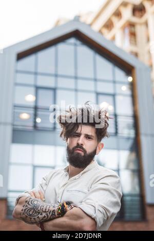 Uomo hipster in piedi con le braccia incrociate contro la costruzione in città Foto Stock