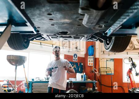 Giovane meccanico che tiene l'attrezzatura guardando l'auto mentre è in piedi in officina di riparazione auto Foto Stock