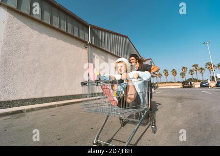 Giovane uomo che spinge la ragazza seduta nel carrello fuori supermercato Foto Stock