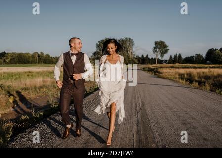 Felice sposa e sposo che corrono su strada durante la giornata di sole Foto Stock