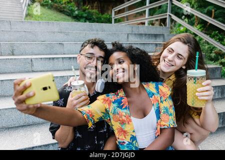 Amici che prendono selfie su smartphone mentre sono seduti in pubblico parcheggio Foto Stock