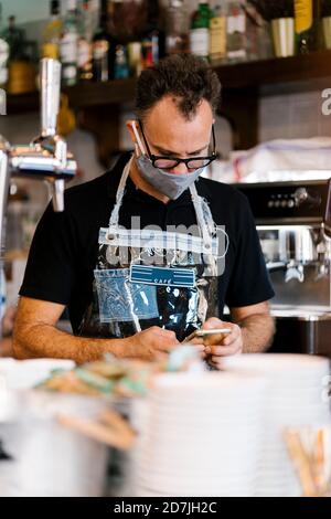 Barista maschile in maschera protettiva con smartphone AT cafe durante il COVID-19 Foto Stock