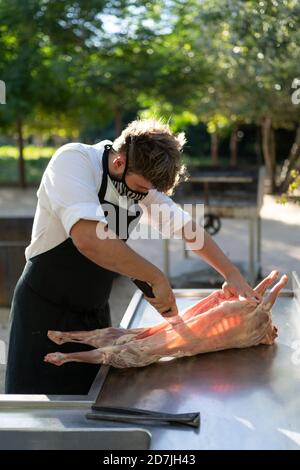 Chef maschile indossare maschera taglio carne di capra sul tavolo mentre in piedi in frutteto Foto Stock