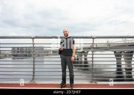 Sorridente giovane uomo con pattini in linea utilizzando smartphone mentre in piedi sul ponte Foto Stock