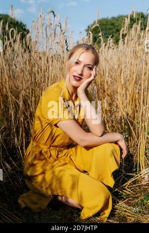 Bella donna bionda accovacciata contro le colture su campo in sole giorno Foto Stock